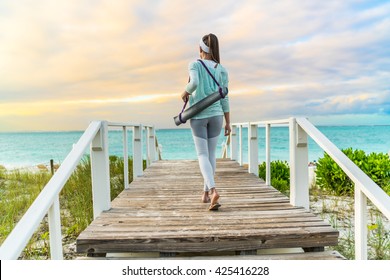 Fitness Woman Walking With Yoga Mat On Beach Going To Outdoor Meditation Class At Sunset. Back View Of Fit Athlete In Activewear Fashion Leggings And Turquoise Hoodie. Healthy Active Lifestyle.