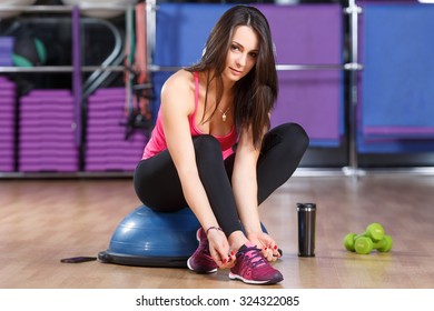 Fitness Woman, Wearing In Shirt, Sneakers And Black Leggings, Sitting On A Bosu Ball And Tying Her Shoe Laces. Cup, Dumbbells And Smart Phone Lying On The Floor, In The Gym, Full Body