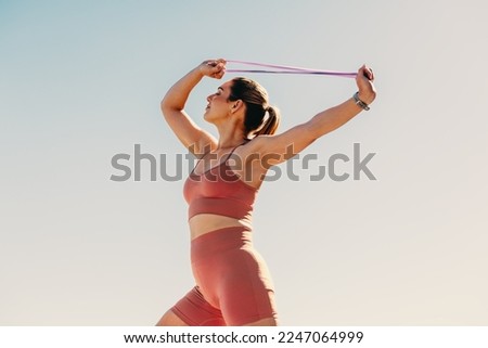Fitness woman working out with resistance bands outdoors. Active sports woman doing strength training on her arm muscles. Woman exercising against the sky.