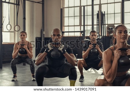 Fitness women and determined men exercising with kettlebell at gym. Group of young people doing a kettle bell exercise with squatting. Multiethnic group doing crouch exercise while holding weight.