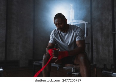 Fitness workout in gym. African American man fighter preparing for fight wrapping hands with red boxing wraps sports protective bandages in gym. Strong man ready for fight boxing sparring training - Powered by Shutterstock