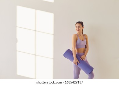 Fitness yoga exercise workout woman. A happy woman with a yoga mat in her hands stands against a white wall in a bright room.