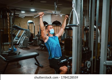 A Fitness Young Caucasian Man With A Face Mask Is Exercising In The Gym And Using Machine For Arm. Protection Against COVID - 19 Coronavirus