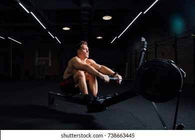 Fitness Young Man Using Rowing Machine In The Gym. Rower Bodybuilder Trains In The Loft Space. Torso In Shorts. Concentrated Motivation.