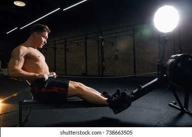 Fitness Young Man Using Rowing Machine In The Gym. Rower Bodybuilder Trains In The Loft Space. Torso In Shorts. Concentrated Motivation.