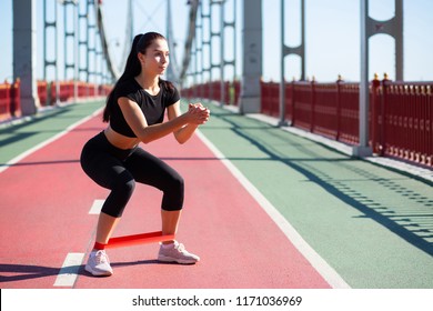 Fitness Young Woman In Sport Apparel Crouching Using Resistance Band While Workout. Space For Text