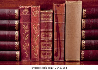 Five antique  books  standing up on a polished wooden shelf supported by other old books on their sides.