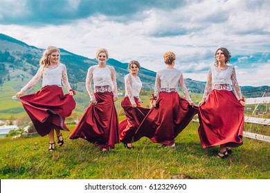 Five Beautiful Bridesmaids In Same Color Red Dress On Wedding Day Having Fun And Circling Around Dresses Behind Mountains And Blue Sky. Funny Girls.