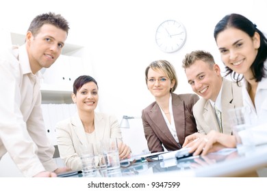 Five business colleagues sitting around table and working together, looking at camera, smiling.