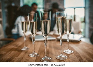 Five Champagne Glasses Standing On Negotiation Table At Business Event, Team Of Coworkers Celebrating Agreement Or Successful Project In Office On Blurred Background, Toned Image
