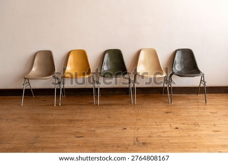 Five colorful plastic chairs lined up against a plain wall on a wooden floor, creating a minimalist and retro aesthetic.
