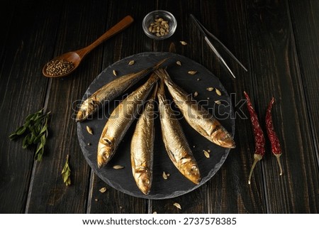Five fish are placed on a round slate plate. Around the plate, there are spices, herbs, and kitchen tools. The setting is dark with a cozy atmosphere as evening approaches.