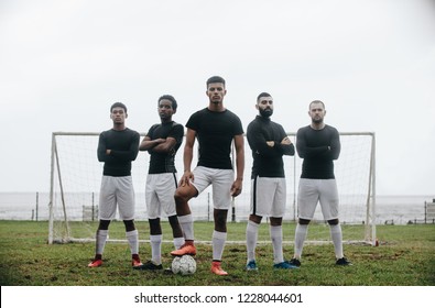 Five Football Players Standing In A Formation In Front Of Goalpost. Soccer Player Standing With One Foot On Ball With Teammates Standing Behind Him With Arms Crossed.