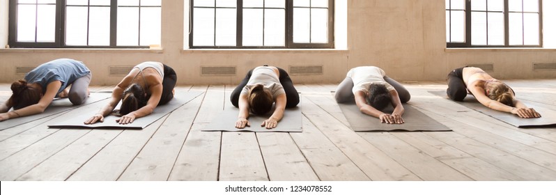 Five girls during yoga session at sport studio. Sportive females lying in row relaxing on wooden floor rubber mats doing Child Pose. Horizontal photography banner for website header. Wellness concept