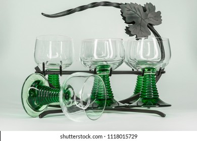 Five Green Wine Glasses With Rippled Green Stems Sitting In A Wrought Iron Rack With One Glass Tipped Over.