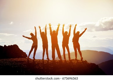 Five happy friends having fun standing with raised arms on mountain top at sunset time