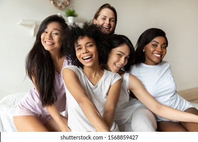 Five happy multiracial young ladies wear sleepwear posing together sitting on bed, smiling diverse brunette girls gathering on slumber party look at camera, portrait, multi ethnic friendship concept