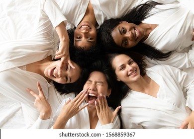 Five happy pretty diverse young girls friends wear robes lying in circle on bed laughing having fun, smiling multi ethnic ladies looking at camera celebrate hen bachelorette party, top view portrait