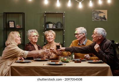 Five Happy Senior Friends With Pieces Of Paper On Foreheads Sitting At Dining Table, Playing Guessing Game And Clinking Wine And Champagne Glasses