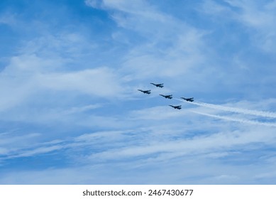 Five jets fly in a V-formation, emitting white smoke trails against a backdrop of a partly cloudy sky.