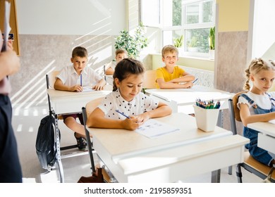 Five Nice Pupils Are Listening Carefully To Their Tutor. Primary School Kids Sitting On Desks And Working In Classroom. Education And School Concept.