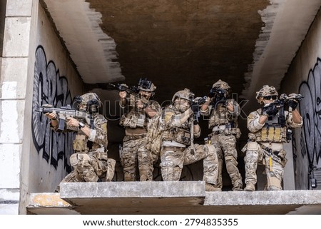 Five operators in full camouflage kit staged on a rooftop ledge in an urban AO. One relays SITREP over comms while another runs digital ISR on a laptop. Others provide 360 security with rifles up. The scene captures a forward command element executing reconnaissance, comms relay, and overwatch in a contested battlespace.