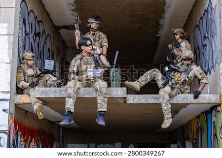 Five operators in full camouflage kit staged on a rooftop ledge in an urban AO. One relays SITREP over comms while another runs digital ISR on a laptop. Others provide 360 security with rifles up. The scene captures a forward command element executing reconnaissance, comms relay, and overwatch in a contested battlespace.