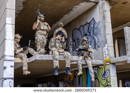 Five operators in full camouflage kit staged on a rooftop ledge in an urban AO. One relays SITREP over comms while another runs digital ISR on a laptop. Others provide 360 security with rifles up. The scene captures a forward command element executing reconnaissance, comms relay, and overwatch in a contested battlespace.