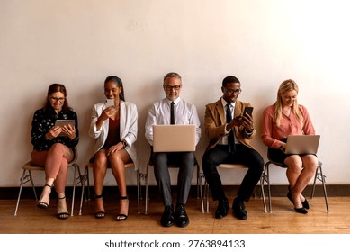 Five professionals sitting in a row, using laptops and smartphones, dressed in business attire, against a plain wall.