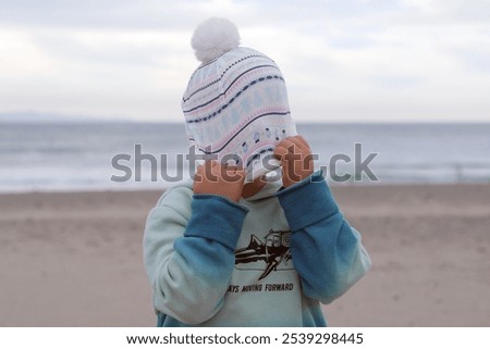 five, six, or seven year old school aged child stands on beach pulling their winter or Christmas themed stocking cap down to hide their face or to be silly