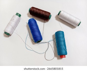 Five spools of sewing thread in blue, turquoise, brown, and white are scattered on a white surface. Loose threads from the spools are tangled in the center, creating a colorful, crafty scene.