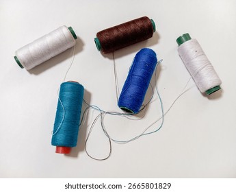 Five spools of sewing thread in blue, turquoise, brown, and white are scattered on a white surface. Loose threads from the spools are tangled in the center, creating a colorful, crafty scene.