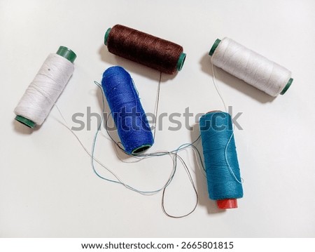 Five spools of sewing thread in blue, turquoise, brown, and white are scattered on a white surface. Loose threads from the spools are tangled in the center, creating a colorful, crafty scene.