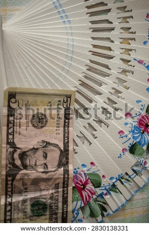 A five US dollar bill placed next to a decorated traditional hand fan, symbolizing financial cooling, inflation, economy, or summer spending concepts.
