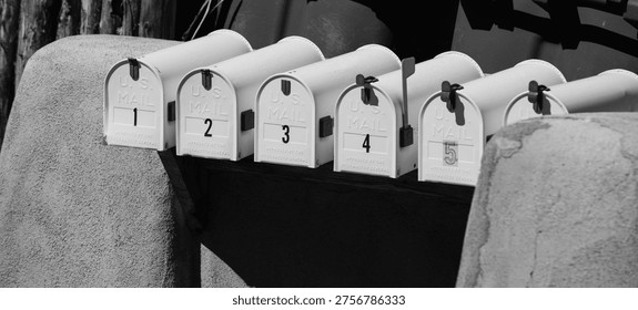 five white mailboxes numbered one through five situated between two adobe style posts all closed showing their numbers horizontal black and white image representing the postal service and snail mail
