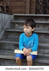 A Five-year-old Dark-haired Boy Sits On A Wooden Porch. He Is Holding A Box With A Chess Set On His Knees, His Gaze Is Directed To The Left. Close-up, Outdoors, Gray Background. Bright Blue Shorts.