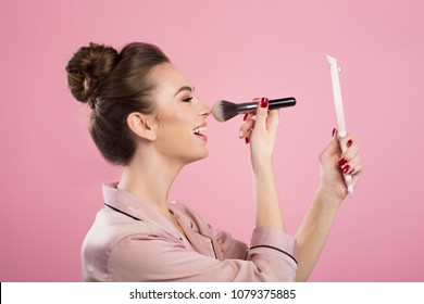 Fix Makeup. Profile Of Cheerful Young Female Is Standing With Hand Mirror And Powdering Her Nose Using Professional Brush. Isolated On Pink Background