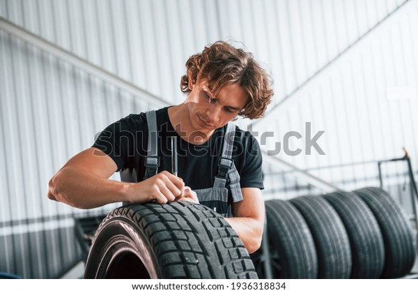 Fixes broken tire. Adult man in grey\
colored uniform works in the automobile\
salon.