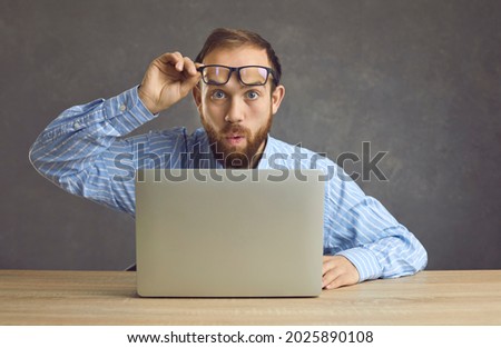 Flabbergasted young man takes off glasses and looks at something with shocked face expression. Surprised worker sitting at desk with laptop computer witnessing unusual situation happening in office Stock foto © 