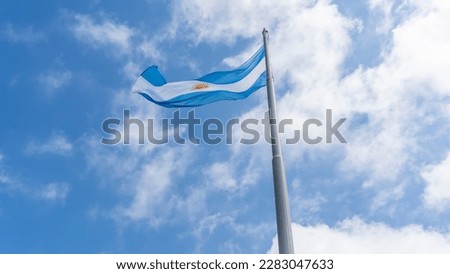 The flag of Argentina against the sky and clouds. A blue-and-white banner with the image of the sun on a high flagpole.