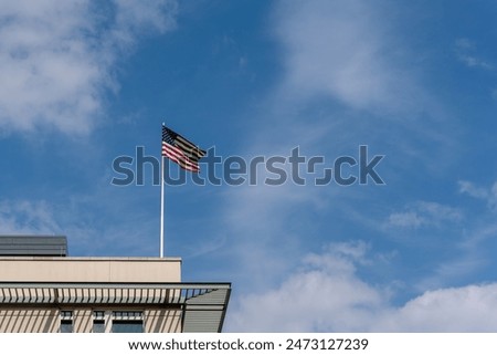 A flag is flying on a pole in front of a building. The sky is blue with some clouds
