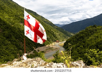 The flag of Georgia waves on a pole atop a rocky outcrop, overlooking a green valley with a winding river, surrounded by forested hills and mountains under a partly cloudy sky - Powered by Shutterstock