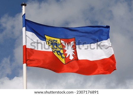 The flag of the German state Schleswig-Holstein with the  state's coat of arms flying in the wind with clouds. The flag is once wrapped around the pole.