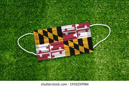 Flag Maryland Placed On A Medical Mask Lies On The Green Grass. Covid-19 Pandemic Concept