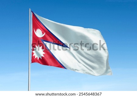 The flag of Nepal features a unique double-pennant design with red, blue, and white colors, showcasing symbols of the sun and moon against a clear blue sky. Flag of Nepal.