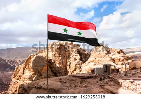 The flag of Syria on a flagpole flutters in the wind against the sky. The Syrian flag is set at a height in the mountains against the backdrop of the nature of the middle East.
