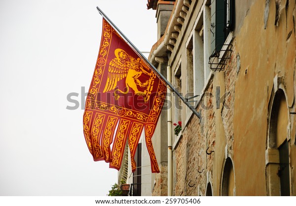 Flag of Venice with winged lion and\
old Venetian building on background. Cloudy winter\
day.
