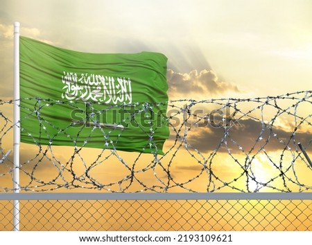 Flagpole with the flag of Saudi Arabia against the sky and behind a fence with barbed wire. The concept of protecting the borders of territories.