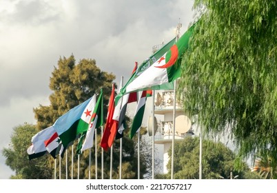 Flags Of Algeria, The Arab League And Arab Countries Flag Waving In The Wind Outside Algiers City With Flagposts Under A Blue Cloudy Sky And Trees In A Sunny Day. Radio Antennas In Background.