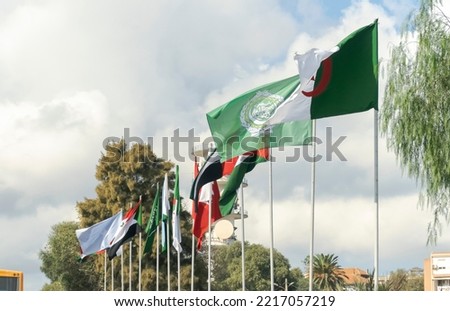 Flags of Algeria, the Arab League and Arab countries flag waving in the wind outside Algiers city with flagposts under a blue cloudy sky and trees in a sunny day.